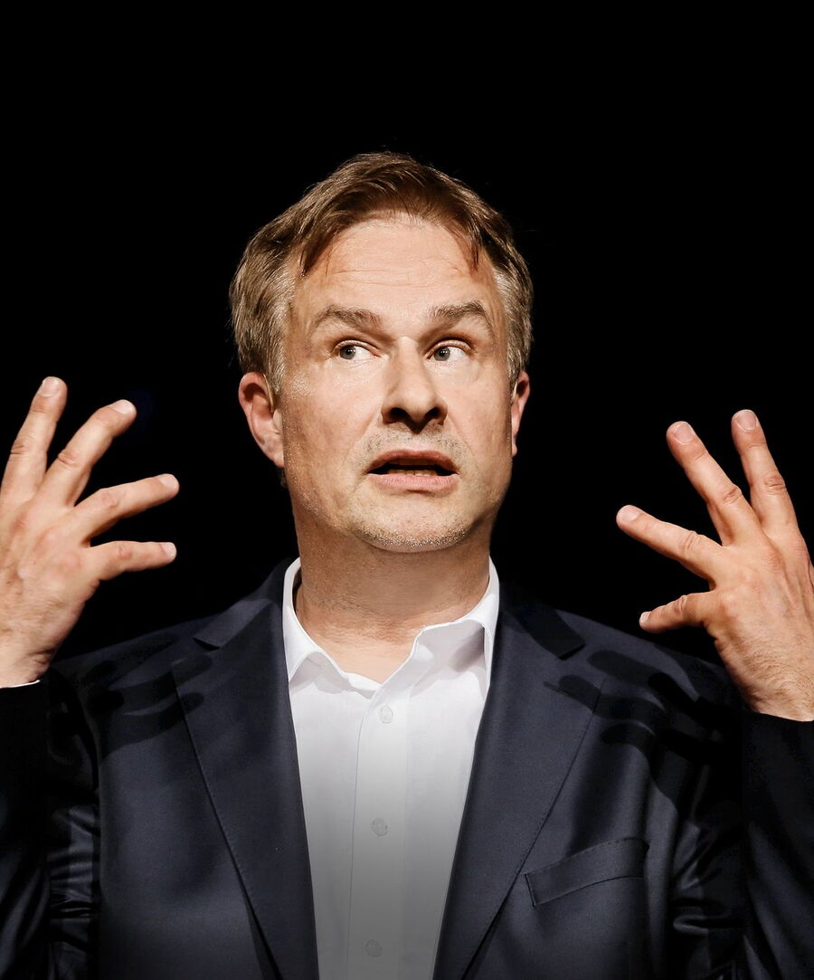 Lars Reichow, ein Mann mit hellbraunem Haar und einem dunkelblauen Anzug mit weißem Hemd, gestikuliert mit beiden Händen nach oben. Er schaut leicht nach oben und scheint mitten im Reden oder Erklären zu sein. Der Hintergrund ist schwarz.