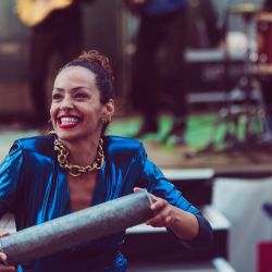 Ayom im blauen glänzenden Kleid, auffälliger, goldener Kette und einem breiten Grinsen. Sie befindet sich vor einer Bühne und trägt etwas in ihren Händen.