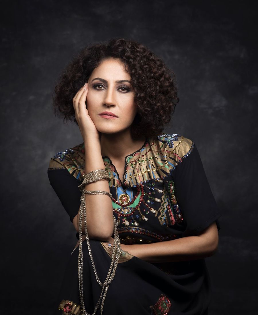 Aynur, eine Frau mit lockigem, dunklem Haar blickt nachdenklich in die Kamera, wobei sie ihre Wange auf ihre Hand stützt. Sie trägt ein kunstvoll verziertes, dunkles Kleid mit goldenen und bunten Details, die reich an Ornamentik sind. Goldene Ketten und S