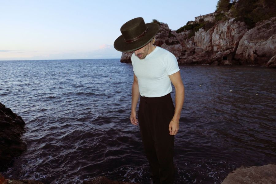 Bonaparte steht am Rand einer felsigen Küste am Meer. Er trägt ein weißes T-Shirt, eine schwarze Hose und einen breiten, dunklen Hut. Der Himmel ist hellblau und im Hintergrund sind steile Felsen sowie Vegetation und ein Gebäude zu sehen.