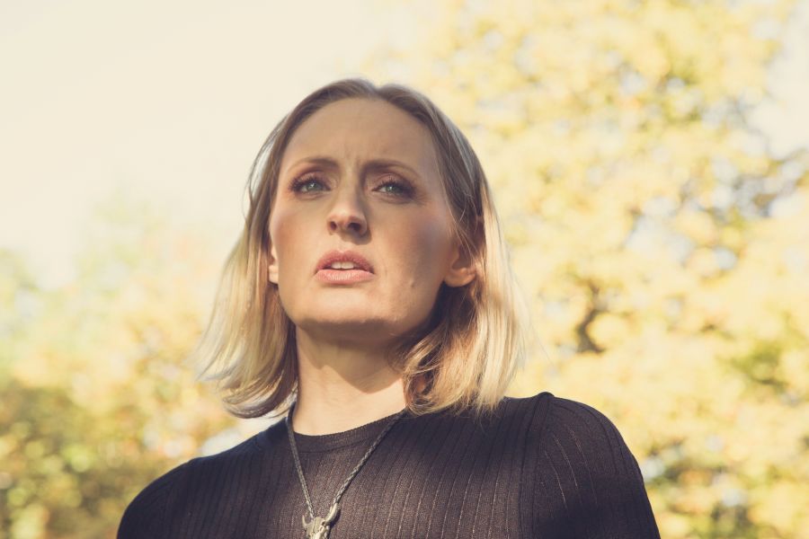 Das Bild zeigt Louisa Lyne ab der Schulter. Der Hintergrund ist in Pastellfarben und unscharf. man kann aber einen blühenden Baum erkennen. Sie selbst trägt ein schwarzes Oberteil, silberne Kette und hat blonde kurze Haare. Sie schaut in die Ferne.