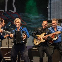 Es sind 4 Musiker auf der Bühne abgebildet, sie alle sind am lachen und musizieren. Das ganze Bild strahlt eine fröhliche, lebendige Atmosphäre aus.