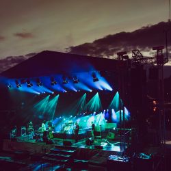 Die Hafensommer Bühne in der Dämmerung. Sie ist blau und grün beleuchet. Das Bild ist sehr dunkel. Auf der Bühne steht eine Band mit 6 Musikern.