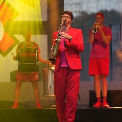 Das Bild zeigt drei Männer in roter Kleidung mit Instrumenten. Einer mit Trommel, einer mit Saxophon und der dritte mit Trompete.