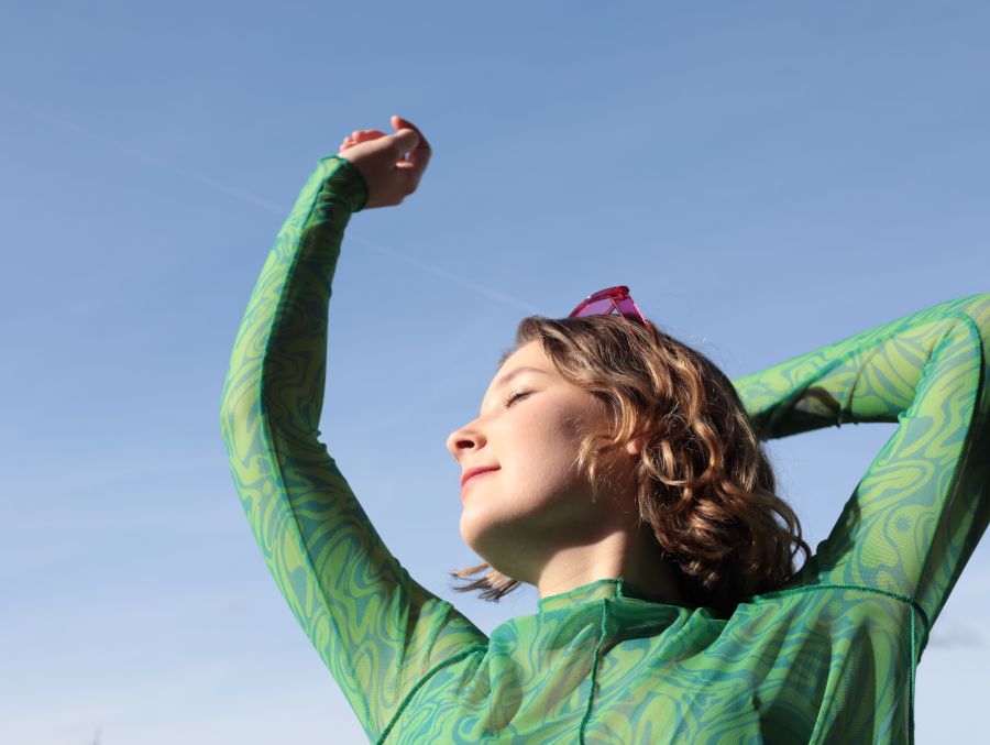 Das Bild zeigt Carla Ahad. Es wurde leicht von unten aufgenommen. Sie steht unter einem wolkenlosen blauen Himmel. Ihr Kopf ist zur Seite gedreht und sie hat die Augen geschlossen, als würde sie die Sonne genießen. Sie trägt ein grünes Oberteil.