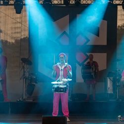 Die Band ist bereit zum spielen. alle der drei Männer welche durch Scheinwerfer hervorgehoben werden halten Drumsticks in ihren Händen, welche sie gerade noch oben strecken.