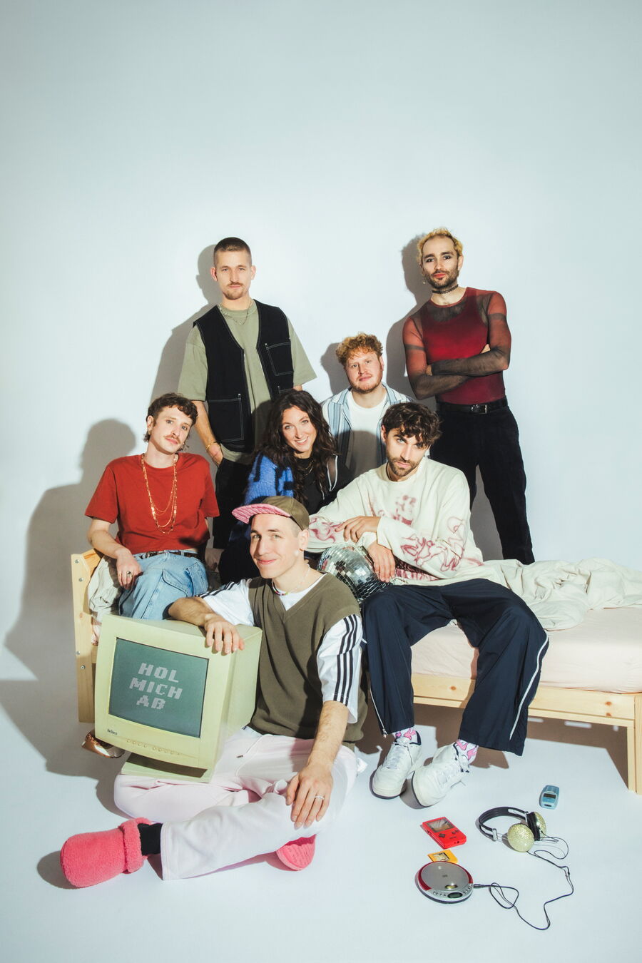 Die Bandmitglieder der Band Make a Move posieren zusammen auf, bzw. bei einem Bett. Vier sitzen auf dem Bett, einer davor. Zwei stehen dahinter. Sie tragen bunte Kleidung und lächeln. Auf dem Schoß des Sitzenden liegt ein Röhrenfernseher.