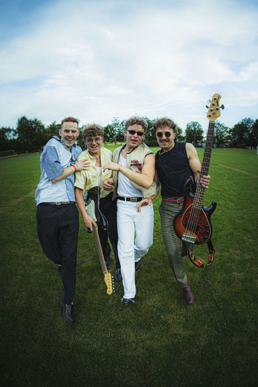 Das Ganzkörperfoto der Band Udo West zeigt die Mitglieder Auf die Kamera zu rennend. Zwei haben Gitarren dabei. Alle haben blonde Locken und zwei tragen eine Sonnenbrille. Sie tragen helle Kleidung und teils gemustert. Das Bild wirkt dynamisch und freundl