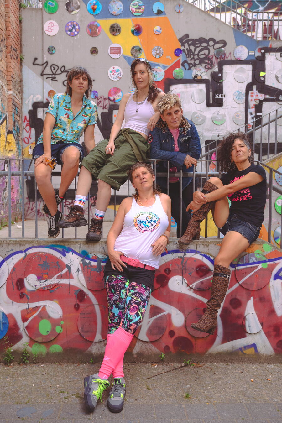Das Bild zeigt die Bandmitglieder von Kumbia Queer vor einer buten Wand, mit einem Geländer voll mit Tags und Graffiti. Sie tragen bunte moderne Kleidung und haben alternative Haarschnitte. Zwei stehen vor dem Geländer und die anderen drei dahinter.