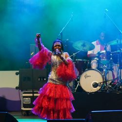 Sängerin in aufälligem rot-goldenen Kleid beim singen mit dem linken arm in der Höhe als würde sie tanzen.