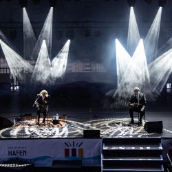 Zwei Musiker, welche mit etwas Abstand auf Stühlen sitzen. Es ist dunkel und sie werden jeweil von Scheinwerfern beleuchtet, die dann auch noch ein Muster auf den Boden werfen.