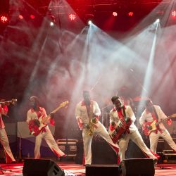 Das Bild zeigt eine Band von 6 personen alle spielen ein Instrument und sie stehen auf einer Bühne. 4 Männer sind in der gleichen Pose, un welcher sie auf dem rechten Bein stehen und das Linke in die Luft heben.