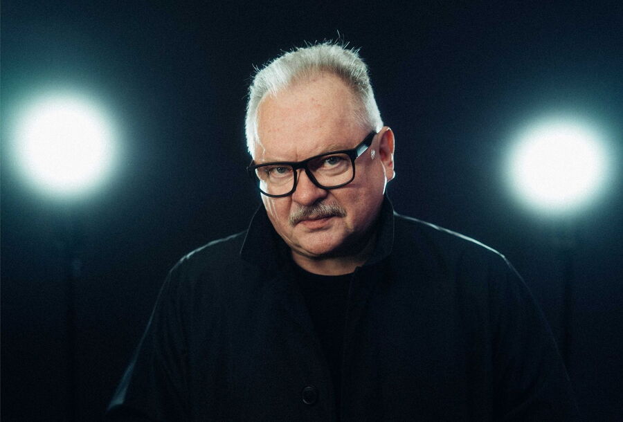 Ein Porträt von Heinz Rudolf Kunze, ein Mann mit kurzen, grauen Haaren und einem Schnurrbart. Er trägt eine schwarze Brille und eine schwarze Jacke. Der Kopf ist leicht geneigt und er blickt direkt in die Kamera. Der Hintergrund ist dunkel, mit zwei helle
