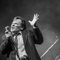 Das schwarz-weiß Foto zeigt einen Sänger mit Mikrofon in der Hand, er hält seinen linken Arm auf Kophöhe, die Finger sind gespreizt.