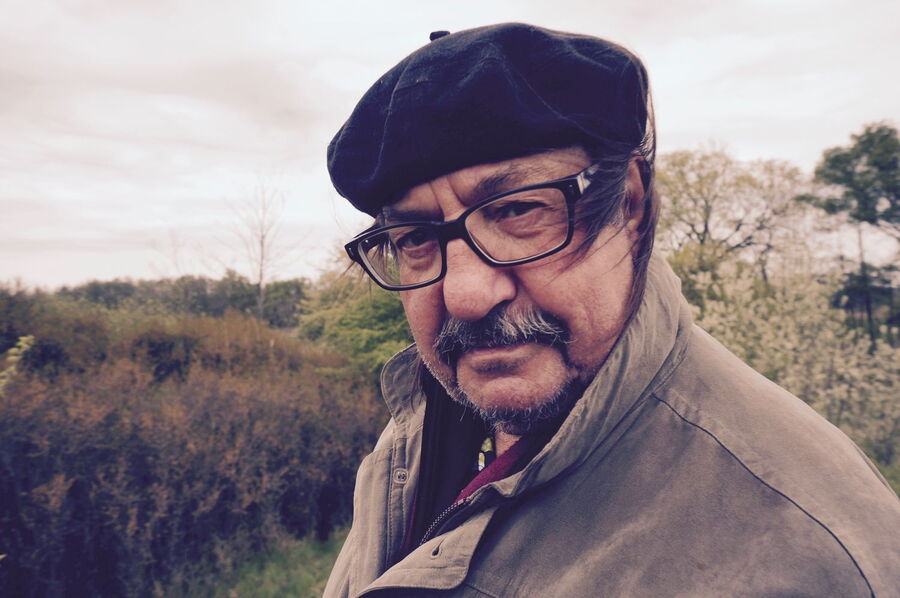 Manfred Maurenbrecher, ein Mann mit Schnurrbart und Brille steht in einer natürlichen Umgebung mit Büschen und Bäumen im Hintergrund. Er trägt eine dunkle Baskenmütze, eine beige Jacke und schaut direkt in die Kamera. Der Himmel ist bewölkt.