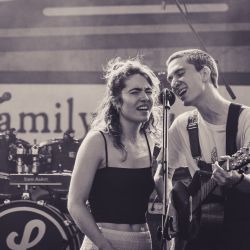 Das Bild zeigt zwei Musiker, die sich ein Mikrofon teilen. Links eine Frau und rechts ein Mann mit Gitarre.