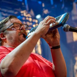 Das Bild zeigt einen Mann im roten Oberteil, welcher auf einer Melodica spielt.