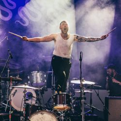 Der Schlagzeuger steht auf seinem Stuhl, er hat beide Arme mit seinen Drumsticks zur Seite ausgestreckt.