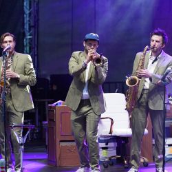 Drei Männer, welche alle den gleichen grünen Anzug tragen. Die beiden äußeren spielen Saxophon der in der Mitte Trompete.