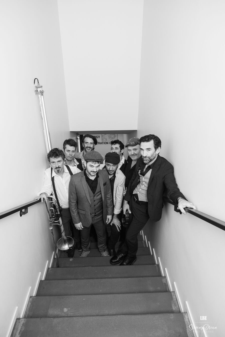 Das Bild zeigt die Mitglieder des Babylon Circus, wie sie auf einer Treppe stehe. Das Bild ist in schwarz-weiß. Die Kamera steht oben auf der Treppe und die Mitglieder unten. Das heißt das Foto ist aus einer leichten Vogelperspektive aufgenommen worden.